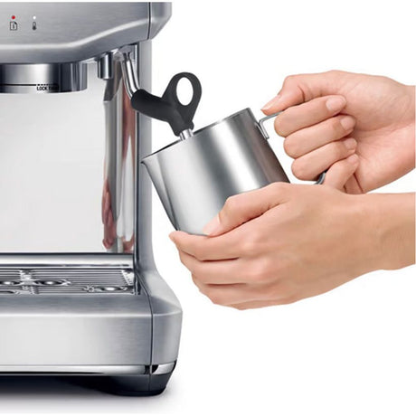 Hands holding stainless steel milk jug under steam wand of Sage Barista Express Impress bean to cup coffee machine in brushed silver finish with black handle and drip tray