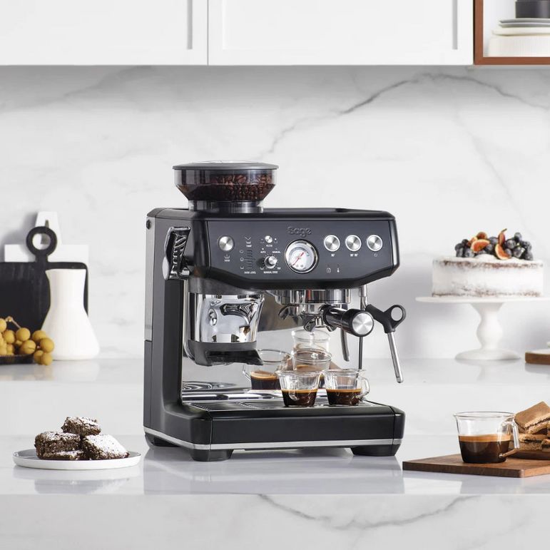 Black Sage Barista Express Impress espresso machine on white marble kitchen countertop surrounded by fresh pastries, coffee cups, and accessories, with white cabinetry and backsplash behind