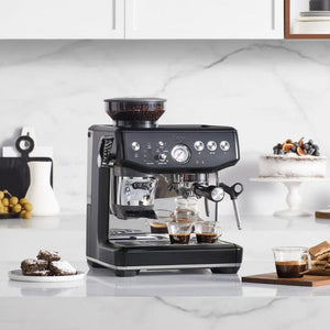 Black Sage Barista Express Impress espresso machine on white marble kitchen countertop surrounded by fresh pastries, coffee cups, and accessories, with white cabinetry and backsplash behind