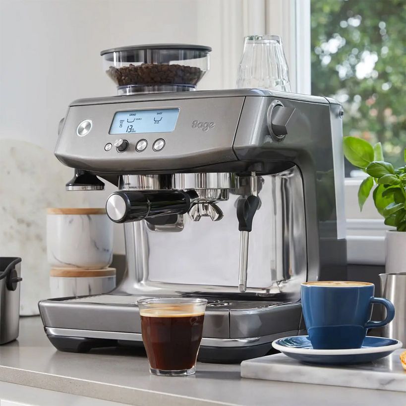 Sage The Barista Pro brushed stainless steel espresso machine on kitchen countertop with integrated grinder, displaying digital display, alongside a cup of fresh espresso and blue ceramic mug with saucer, green plant and window with garden view in background