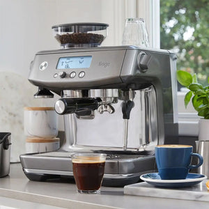Sage The Barista Pro brushed stainless steel espresso machine on kitchen countertop with integrated grinder, displaying digital display, alongside a cup of fresh espresso and blue ceramic mug with saucer, green plant and window with garden view in background