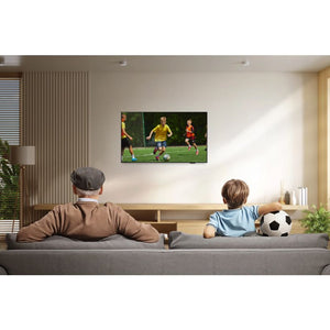 An elderly man and young boy sitting on a gray sofa watching a Samsung F6000 32-inch Full HD Smart TV mounted on a beige wall, displaying a youth soccer match with players in yellow and red jerseys, with a soccer ball on the sofa beside them in a modern living room with wooden furniture and plants