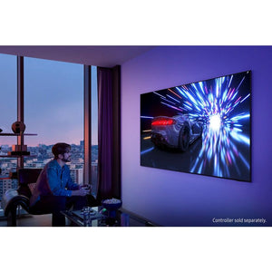 Person sitting in modern high-rise apartment gaming on Samsung 55 inch 4K OLED TV displaying racing game with blue light effects, purple accent lighting on walls, floor-to-ceiling windows with city skyline view at dusk