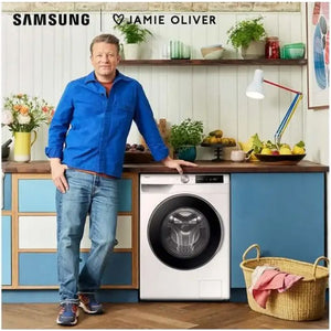 Jamie Oliver standing in a modern kitchen next to a white Samsung Series 6 washing machine with black door, positioned under a wooden countertop with blue cabinetry, surrounded by potted plants, fresh fruit, and a wicker laundry basket