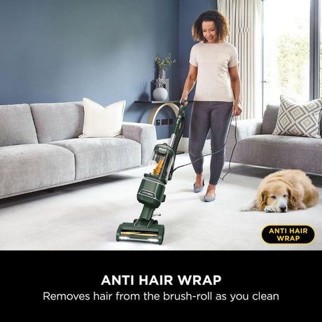 Woman using Shark Anti Hair Wrap upright vacuum cleaner in modern living room with grey sofas, golden retriever dog on light carpet, green and black vacuum with anti hair wrap technology feature highlighted