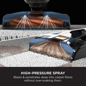 Shark CarpetXpert deep carpet cleaner with navy blue body demonstrating high-pressure spray technology cleaning light gray carpet fibers, showing dual spray nozzles and cleaning brush head in action