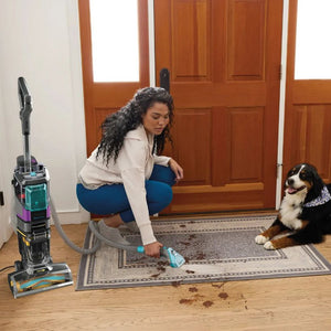 Woman using Shark CarpetXpert HairPro Pet Deep Carpet Cleaner with teal and purple design on hallway mat while brown and white dog watches, wooden doors and hardwood flooring visible in background