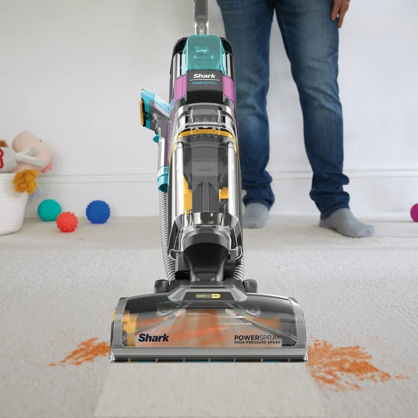 Shark CarpetXpert HairPro Pet deep carpet cleaner in yellow, black and teal being used on beige carpet with orange stains, operated by person wearing blue jeans, with toys scattered nearby
