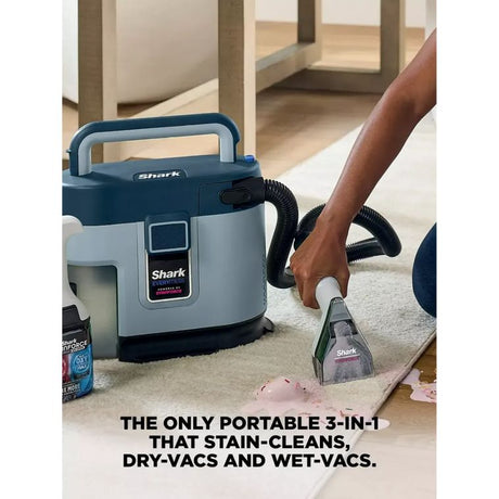 Shark EveryMess 3-in-1 portable spot stain cleaner in blue and grey being used to clean carpet stain, with cleaning solution bottle and hose attachment visible in home setting