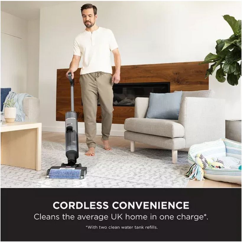 Man using Shark HydroVac cordless vacuum cleaner on light grey carpet in modern living room with beige armchair, wooden headboard, and potted plant, demonstrating cordless convenience for cleaning average UK home with two clean water tank refills