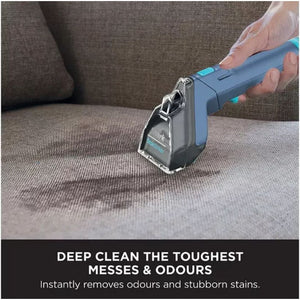 Hand holding blue and black Shark StainStriker portable spot cleaner being used on brown fabric sofa with text below stating Deep Clean The Toughest Messes & Odours Instantly Removes Odours and Stubborn Stains