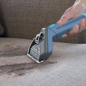 Hand holding a blue and black Shark StainStriker portable stain cleaner with turquoise accents over beige carpet, demonstrating the handheld cleaning device in use