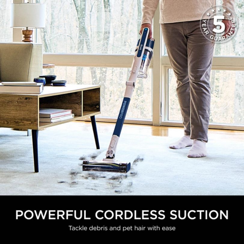 Person in beige sweater and brown pants using a Shark PowerPro cordless stick vacuum with blue and white design to clean light carpet in modern living room with wooden desk and large windows, demonstrating powerful suction collecting pet hair and debris