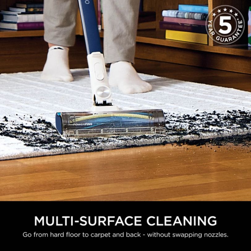 Shark PowerPro cordless stick vacuum demonstrating multi-surface cleaning on hard floor and carpet with white socks visible, showing anti hair wrap technology and floor detect feature, with 5 year guarantee badge