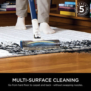 Shark PowerPro cordless stick vacuum demonstrating multi-surface cleaning on hard floor and carpet with white socks visible, showing anti hair wrap technology and floor detect feature, with 5 year guarantee badge