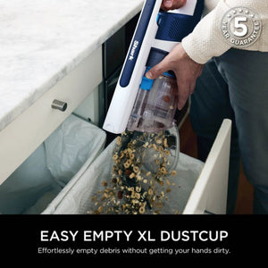 Person holding Shark PowerPro cordless stick vacuum with white and navy blue design, demonstrating easy emptying of XL dustcup filled with debris into white bin, with 5 year guarantee badge visible in top right corner