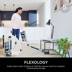 Woman using Shark PowerPro cordless stick vacuum with flexible wand to clean under furniture in modern white kitchen with dark upper cabinets, featuring flexology technology demonstration and 5 year guarantee badge