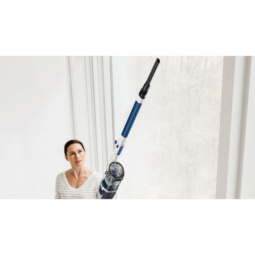 Woman in striped beige top holding extended Shark PowerPro cordless stick vacuum with blue and black body and anti hair wrap technology against white curtained wall