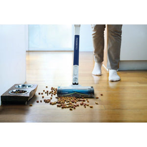 Shark PowerPro cordless stick vacuum in navy blue and silver being used by a person in white socks and tan trousers on wooden floor to clean scattered pet kibble near stainless steel food bowls