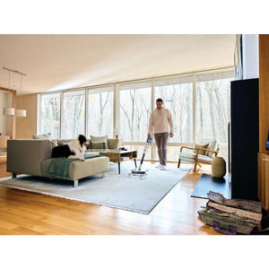 Man using Shark PowerPro cordless stick vacuum in modern living room with beige sofa, wooden flooring, large windows overlooking forest, and woman relaxing on couch