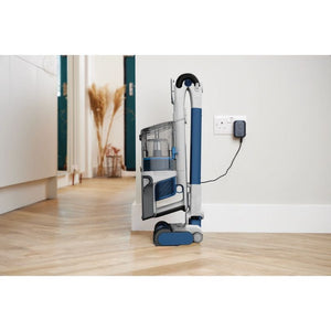Shark PowerPro cordless stick vacuum in white and blue with anti hair wrap technology, displayed on wooden flooring in a modern minimalist home interior, showing the charging dock on the white wall behind it