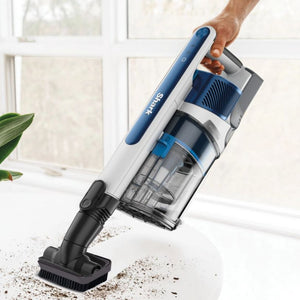 Shark PowerPro cordless stick vacuum cleaner in blue and white being held at an angle over scattered debris on a white floor, with transparent dust cup visible and windows in soft focus background