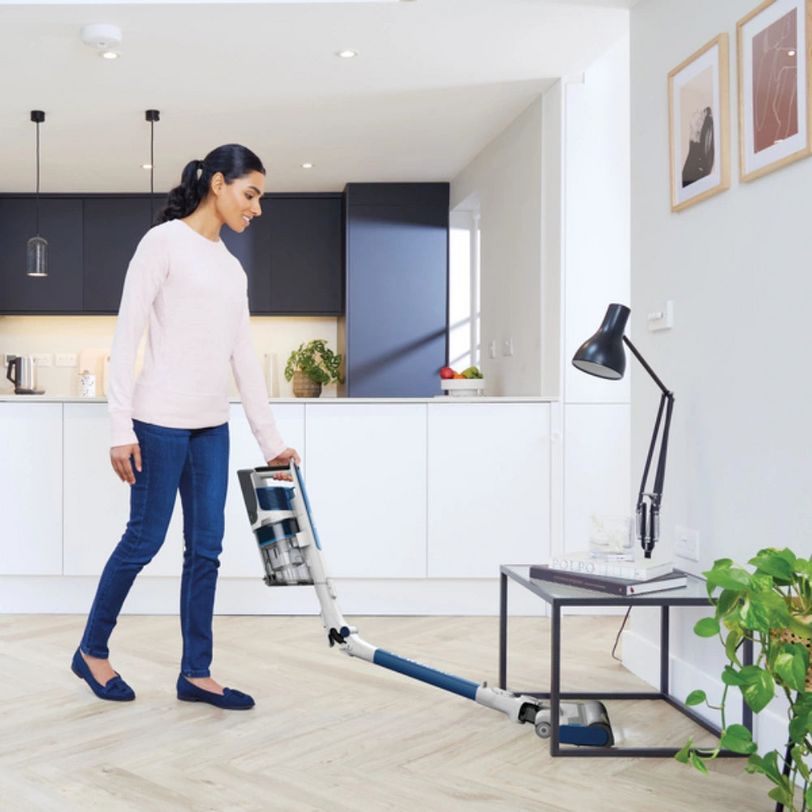 Woman using Shark PowerPro cordless stick vacuum on light wooden flooring in modern kitchen with white cabinetry and black upper units, demonstrating cleaning capability in contemporary home setting
