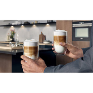 Person holding two tall glass cups of layered cappuccino with white foam in a modern kitchen with Siemens EQ500 black automatic coffee machine in background