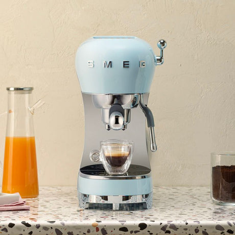 Smeg 50's retro-style espresso pump coffee machine in pastel blue with chrome accents, dispensing dark espresso into a double-walled glass cup, positioned on a speckled countertop with orange juice and dark beverage in glasses beside it