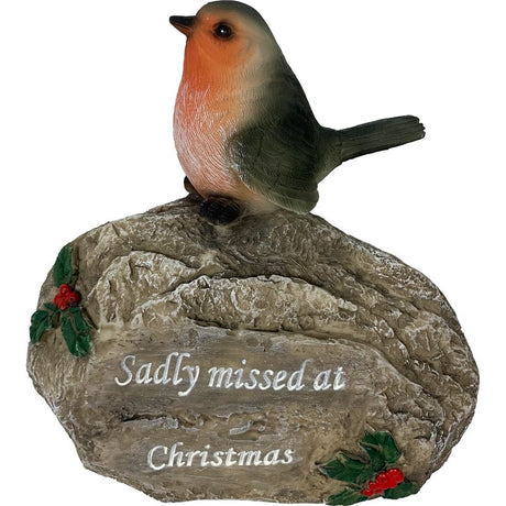 Ceramic robin bird figurine perched on textured grey stone memorial plaque with white engraved text reading Sadly missed at Christmas, decorated with holly sprigs and red berries, isolated on white background