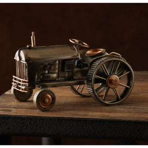 Vintage-style metal tractor ornament with bronze and brass patina finish, featuring detailed wheels, steering wheel, and exhaust pipe, displayed on wooden surface against dark background