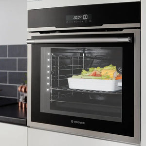 Hoover stainless steel built-in single oven with black glass door displaying vegetables cooking inside, digital display showing 200 degrees, LED interior lighting, and chrome handle in modern kitchen with dark tile backsplash