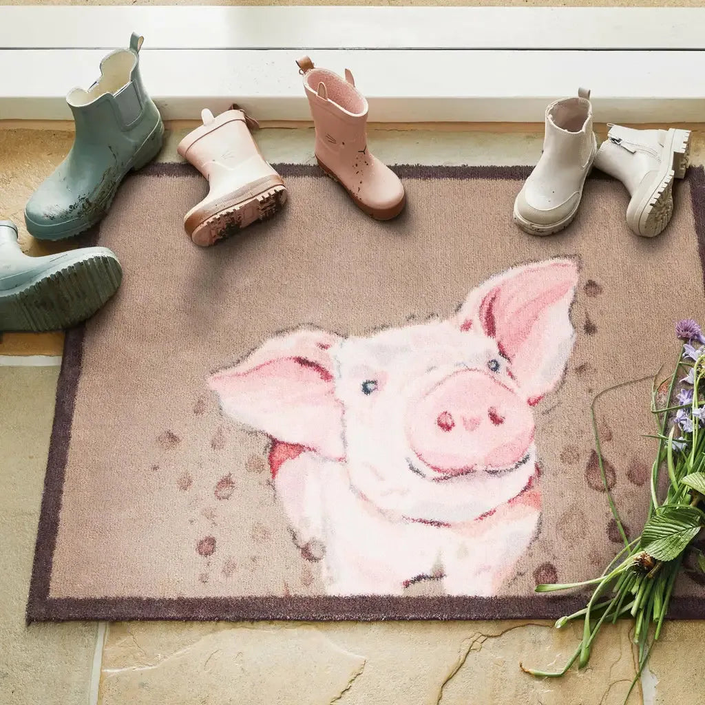 Hug Rug doormat featuring a cheerful pink pig design on a beige and brown woven surface, displayed with children's wellington boots in green, pink, and white arranged above it, with fresh flowers and green plants visible at the side