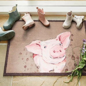 Hug Rug doormat featuring a cheerful pink pig design on a beige and brown woven surface, displayed with children's wellington boots in green, pink, and white arranged above it, with fresh flowers and green plants visible at the side
