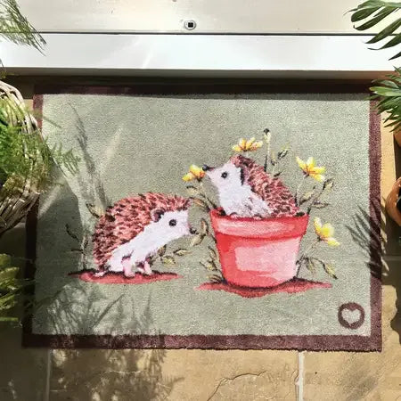 Hug Rug doormat featuring two pink hedgehogs with yellow flowers and a red potted plant on sage green background, 65cm x 85cm size displayed outdoors with plants visible at edges