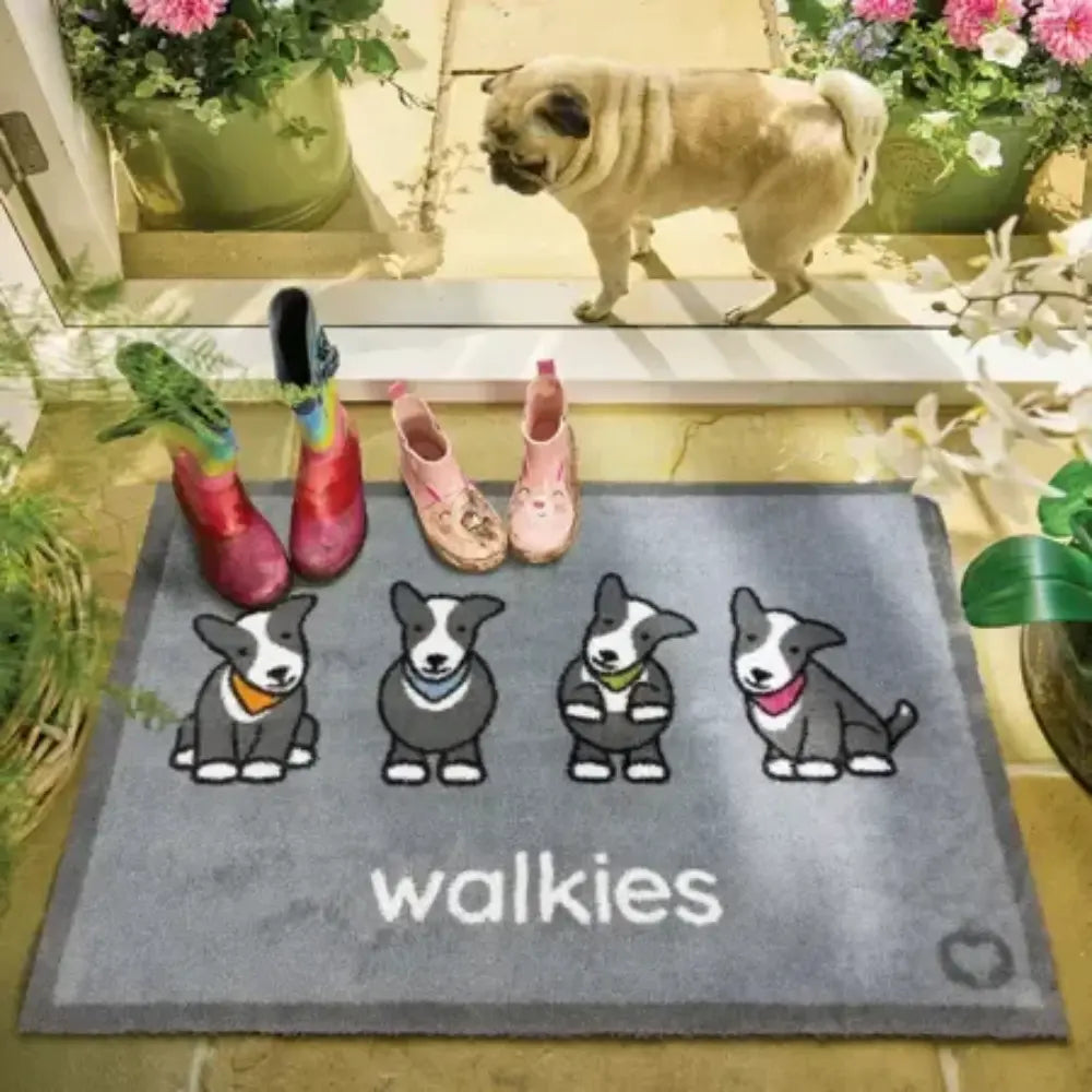 Gray doormat with four illustrated white and tan dogs wearing colored bandanas and the text 'walkies' below, displayed on a pale yellow floor surrounded by potted pink and white flowers, with colorful children's boots and a tan pug visible above the mat