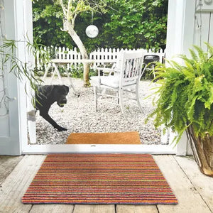 Hug Rug doormat 50cm x 75cm in candy stripe bright pattern placed at entrance of open patio doors overlooking garden with potted fern and black dog on gravel patio