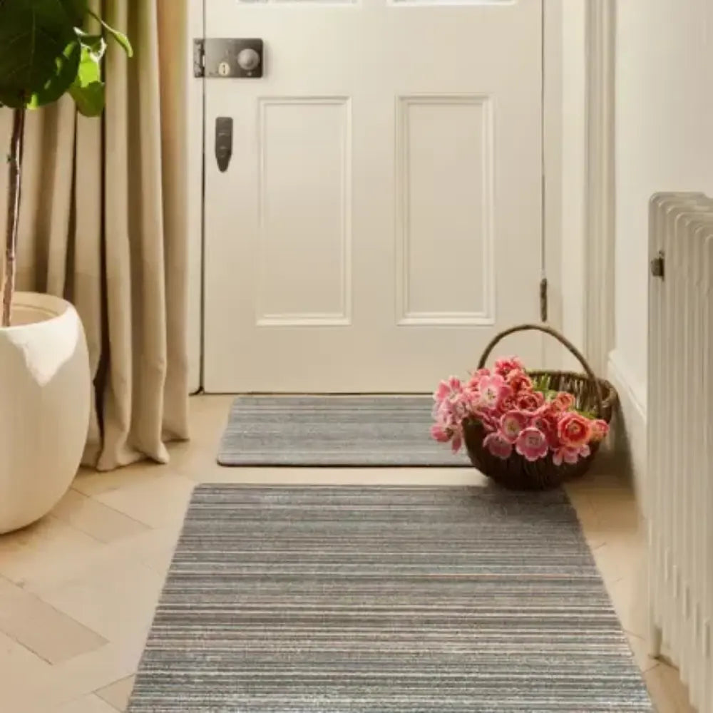 Hug Rug doormat 80cm x 100cm in sand colour placed in a bright entryway with white double doors, grey striped runner mat, pink flowers in woven basket, white planter with greenery, and beige curtains