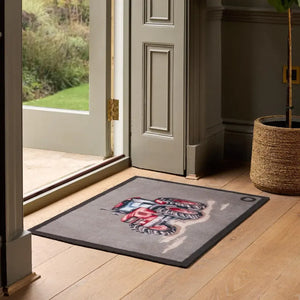 Hug Rug Farmyard Tractor doormat 65cm x 85cm displayed in an entryway showing a red tractor design on grey background with black border, placed on wooden flooring in front of open glass door with garden view
