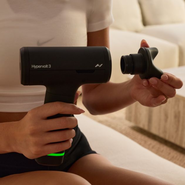 Person holding black Hyperice Hypervolt 3 massage gun with detachable massage head in a bright modern living room setting