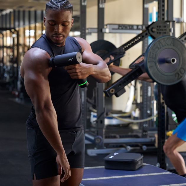 Man in black sleeveless shirt using Hyperice Hypervolt 3 Pro massage gun on arm in modern gym with weightlifting equipment in background