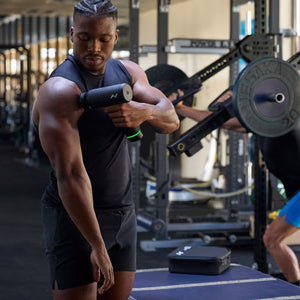 Man in black sleeveless shirt using Hyperice Hypervolt 3 Pro massage gun on arm in modern gym with weightlifting equipment in background