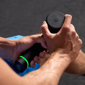 Hands holding black Hyperice Hypervolt 3 Pro massage gun with green accent ring and blue fabric surface visible in background