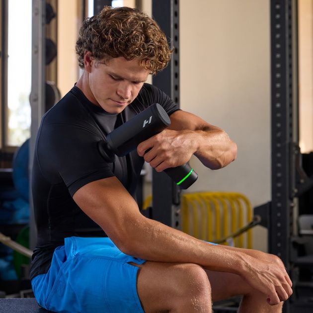Man in black t-shirt and blue shorts using Hyperice Hypervolt 3 Pro massage gun with green accent on his arm in a gym setting