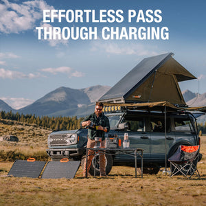 Man standing at camping setup with roof tent and portable solar panel charging devices next to SUV in mountain landscape