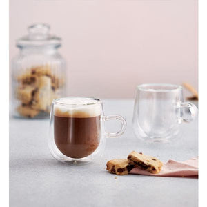 Two clear double-walled glass mugs on a light gray surface, with one mug filled with a dark coffee drink and foam top, the other empty, with cookies and a glass jar blurred in the background.