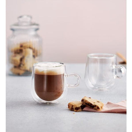 Two clear double-walled glass mugs on a light gray surface, with one mug filled with a dark coffee drink and foam top, the other empty, with cookies and a glass jar blurred in the background.