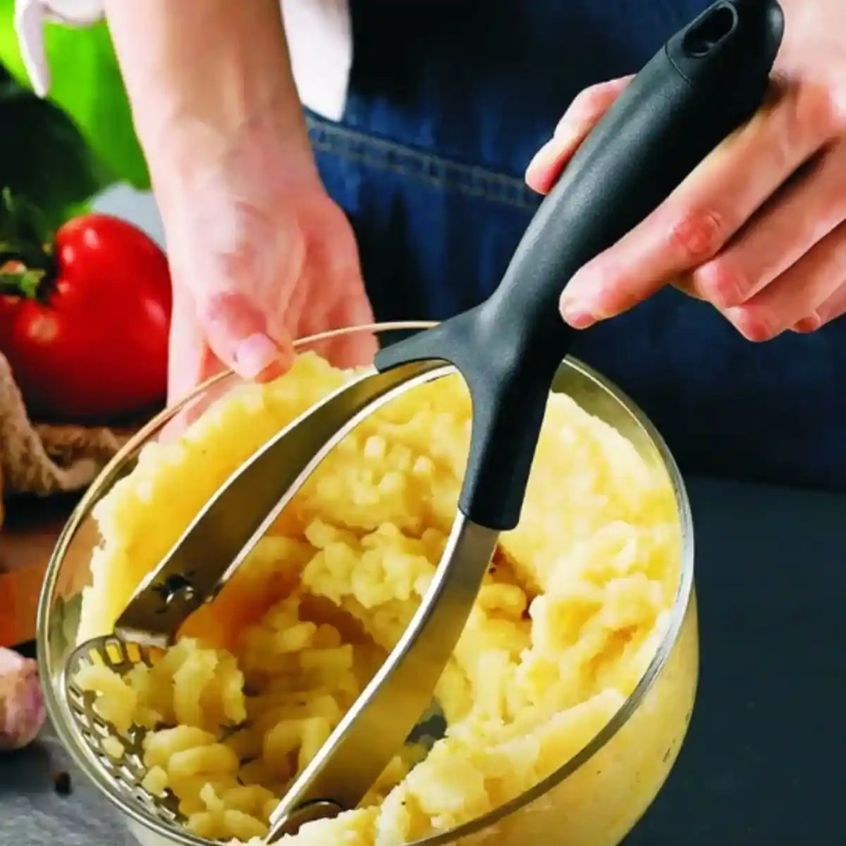 Person using a black-handled folding potato masher to mash cooked diced potatoes in a clear glass bowl, with fresh red tomato and green vegetables visible in the background