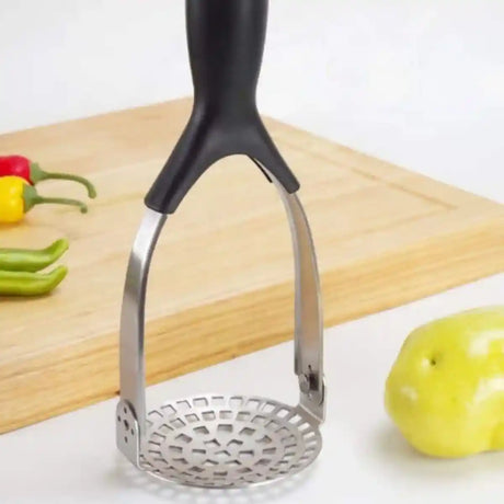 Stainless steel folding potato masher with black handle on wooden cutting board surrounded by fresh vegetables including red pepper, yellow pepper, green pepper slices and green apple