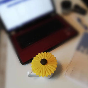 Yellow silicone daisy-shaped cup lid with dark center displayed on a light surface next to a red and black mug, with a smartphone and other items blurred in the background